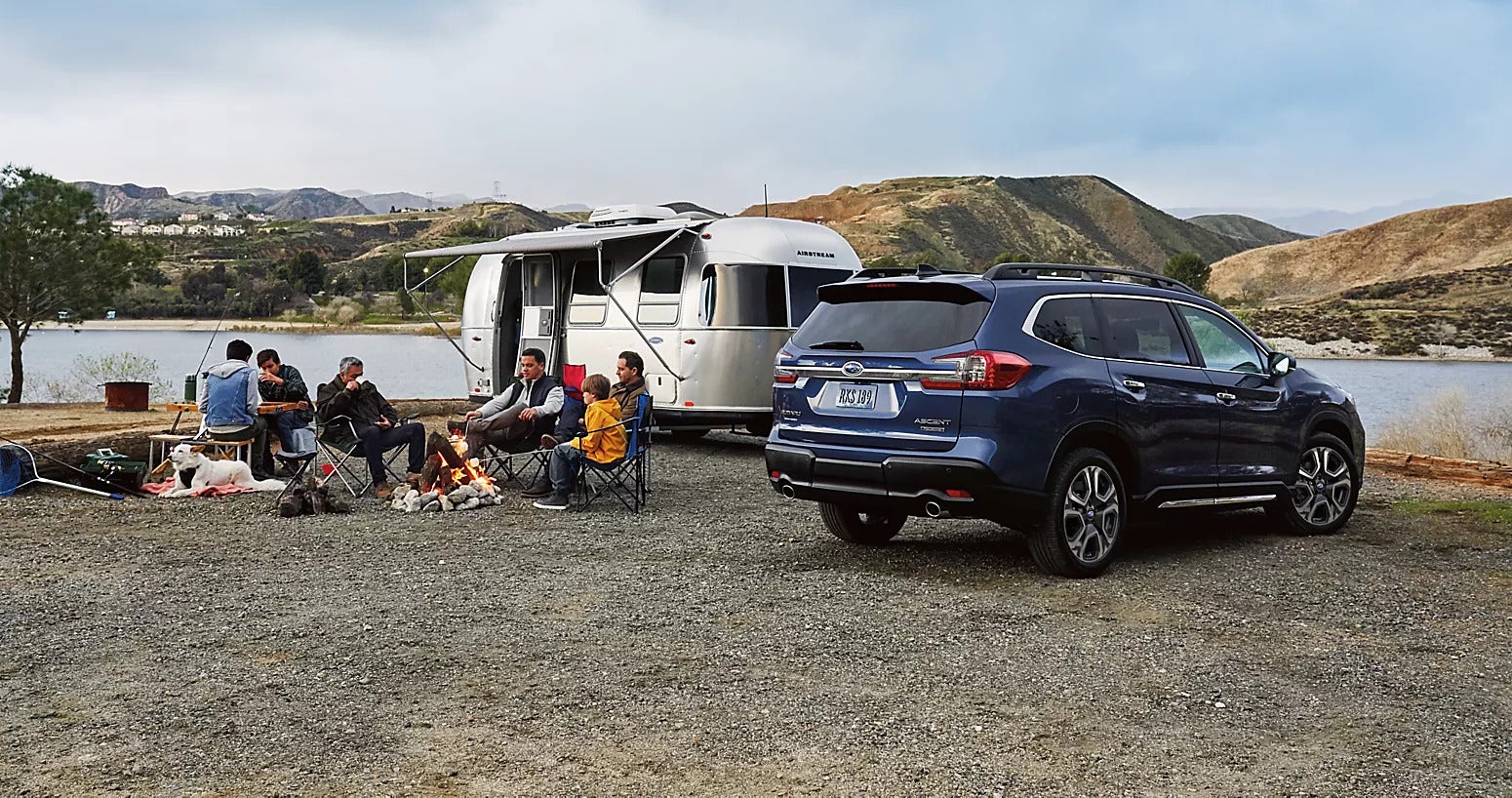 a group of people camping next to a silver camper and a blue subaru ascent near a lake